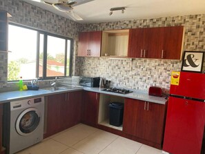 A compact kitchen with wooden cabinets, a washing machine, a gas stove, and a red refrigerator. - Captivating 2-bed Ensuite Apartment in Accra (Accra)