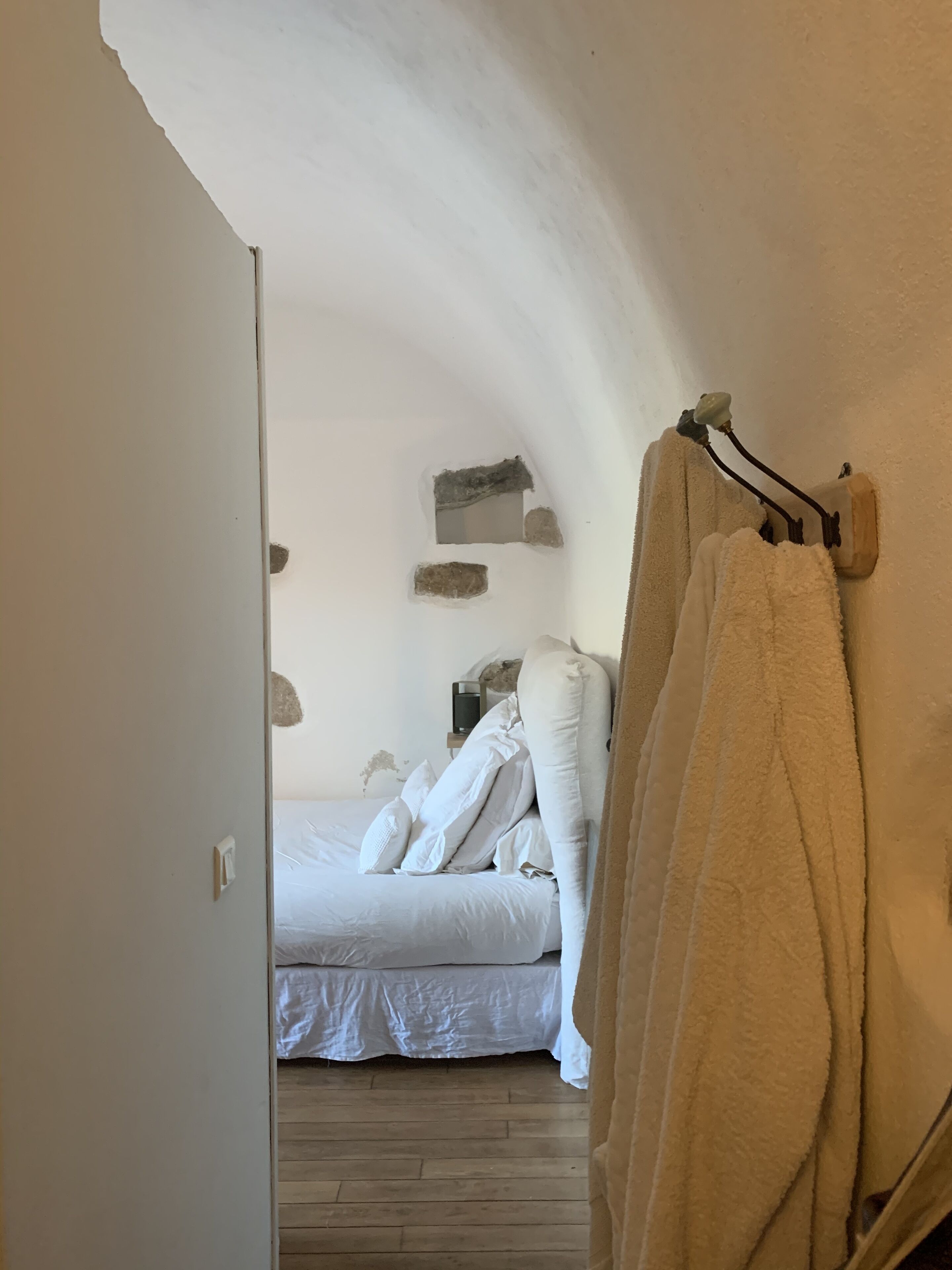 Un dormitorio con cama, ropa de cama blanca, suelo de madera y una toalla colgada de un gancho.