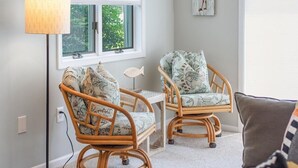 A room with two wicker chairs with patterned cushions, a small table, a lamp, and a window with blinds. - Coastal Chic 2BR Condo w/ Community Pool, Walk to Restaurants (Rehoboth Beach)