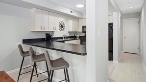 A modern kitchen with a black countertop, white cabinets, and a clock on the wall. There are three bar stools and a refrigerator in the background. - Coastal Chic 2BR Condo w/ Community Pool, Walk to Restaurants (Rehoboth Beach)