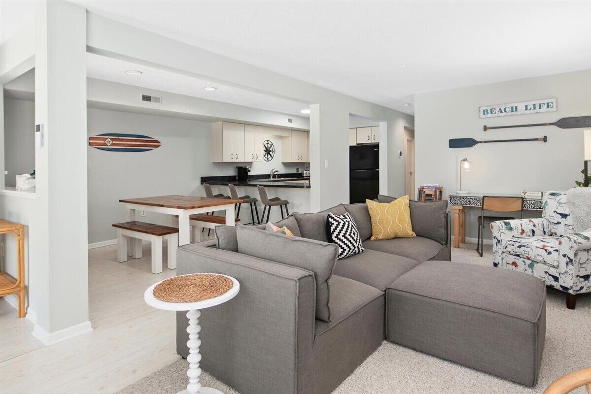 Un salon moderne avec un canapé d’angle gris, un coin repas avec une table et des chaises en bois, et une cuisine en arrière-plan.