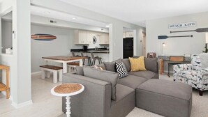 A modern living room with a grey sectional sofa, a dining area with a wooden table and chairs, and a kitchen in the background. - Coastal Chic 2BR Condo w/ Community Pool, Walk to Restaurants (Rehoboth Beach)