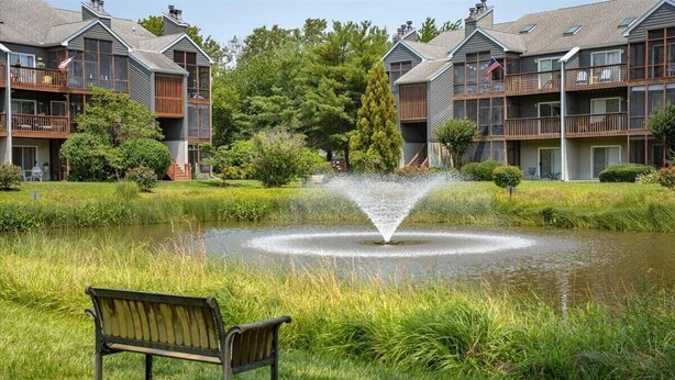 A multi-story building with balconies, a pond with a fountain, and a bench. - Coastal Chic 2BR Condo w/ Community Pool, Walk to Restaurants (Rehoboth Beach)
