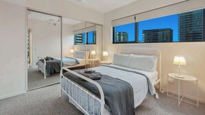Ein Schlafzimmer mit Bett, Nachttischen, Deckenventilator, einem großen Spiegel und Blick auf Gebäude durch das Fenster.