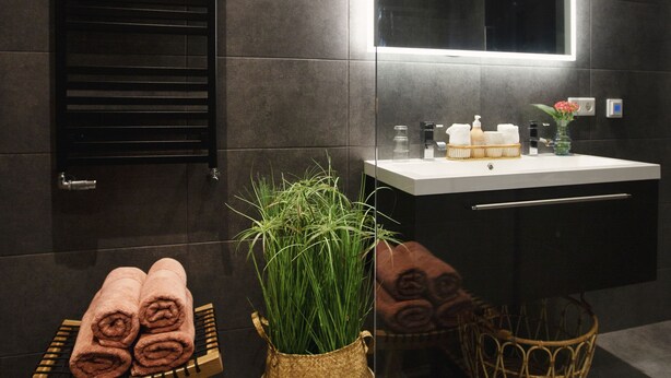 A modern bathroom with a dark tiled wall, a black towel rack, a wooden stool with towels, a large mirror, and a sink with toiletries.