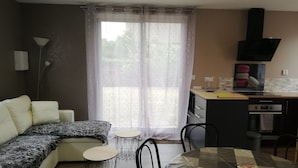 A modern living room with a sofa, a dining table with chairs, a kitchen area with a stove and microwave, and a large window with sheer curtains. - House 42 m2, 200 m from the sandy beach, with enclosed garden and parking. (Portbail)
