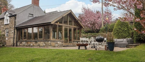 A stone house with a large glass windowed porch, a barbecue area, and a garden with blooming trees.