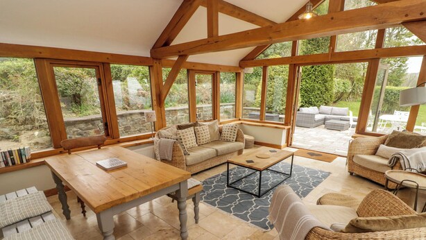 Una habitación acogedora y bien iluminada con una mesa de madera, un sofá y una silla de mimbre. Amplias ventanas ofrecen vistas al jardín.