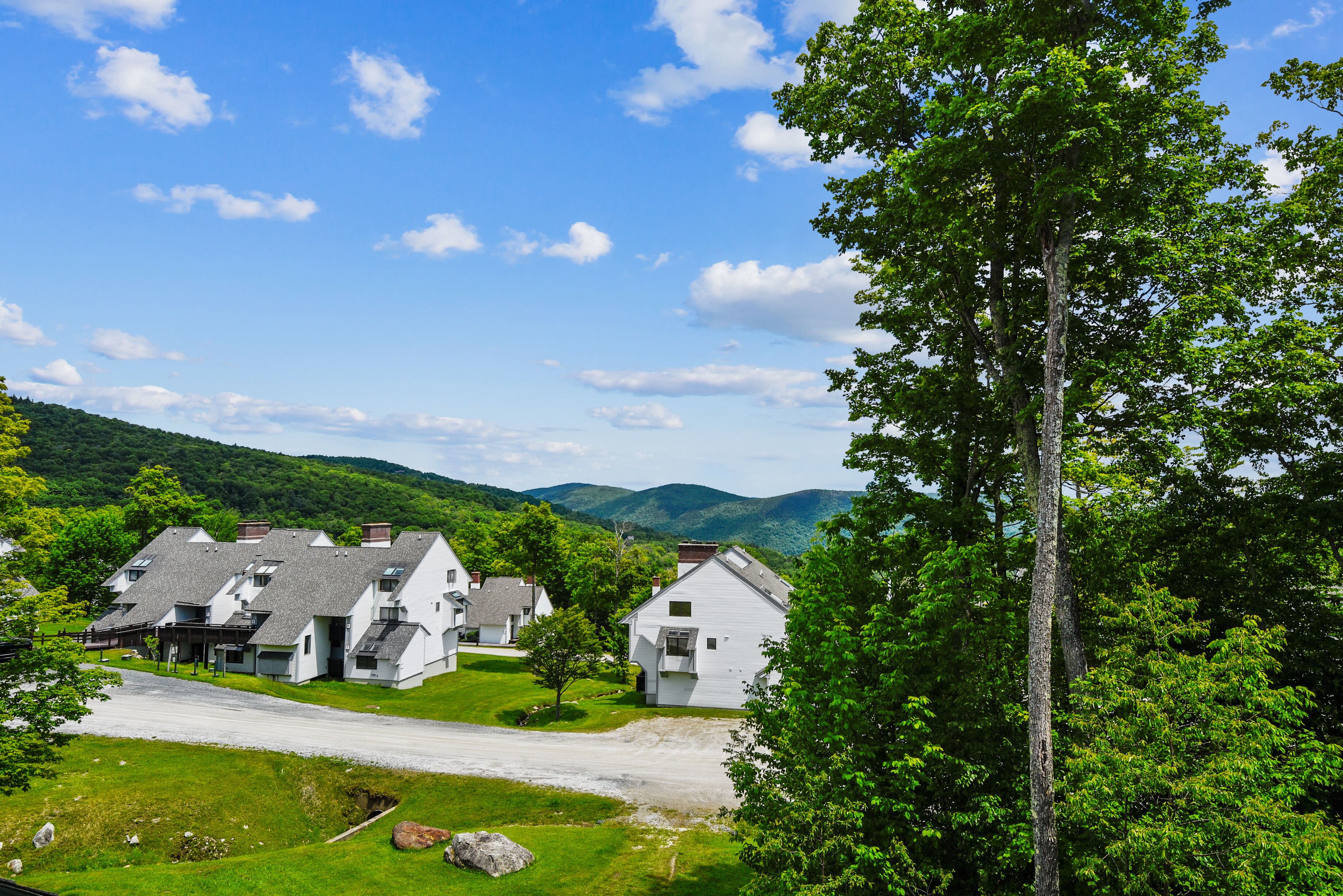A cluster of white houses with sloped roofs, surrounded by lush greenery and mountains in the background.
