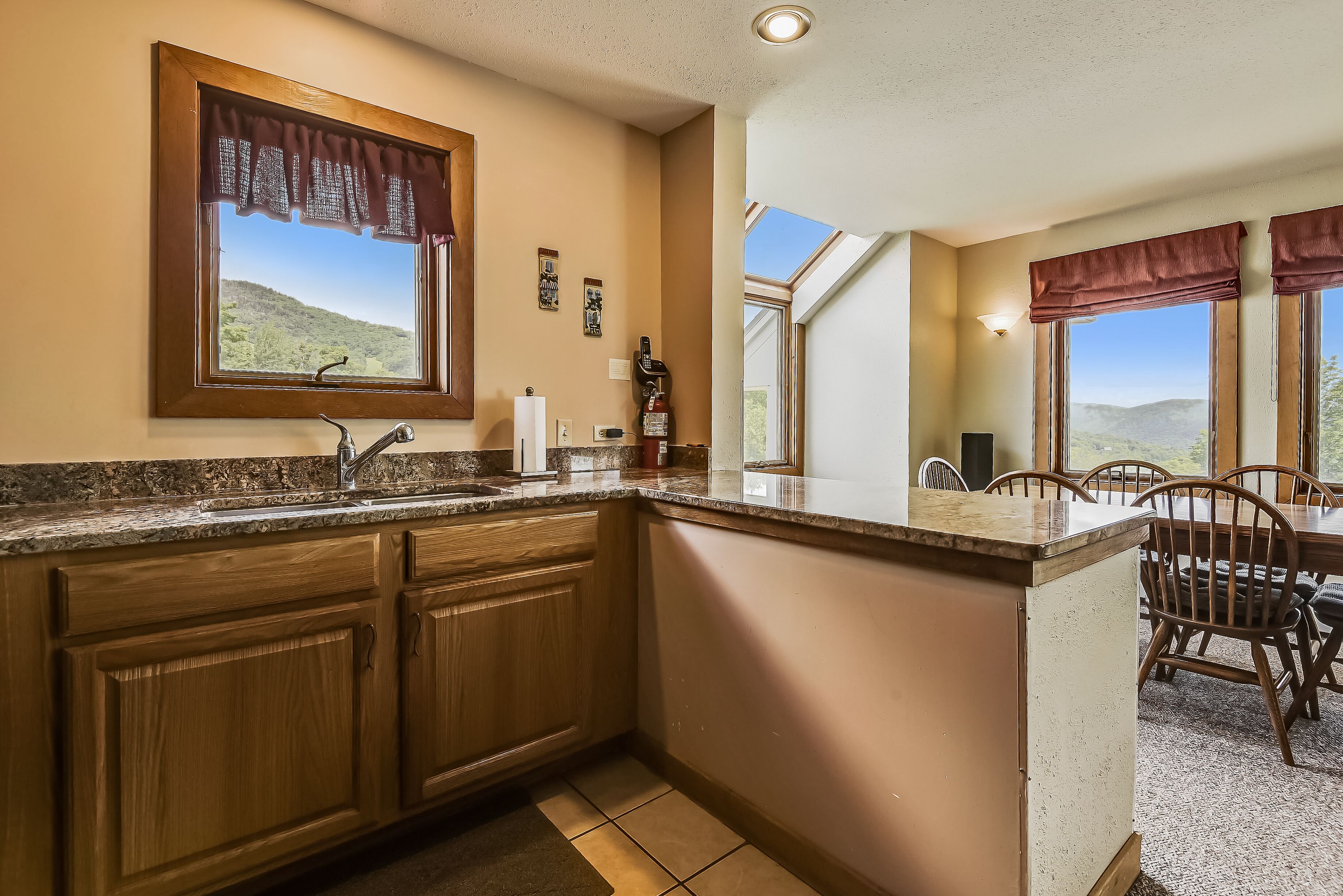 A kitchen with wooden cabinets, a granite countertop, a window with a view of a mountain, and a dining area with chairs.