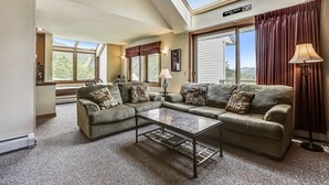 A living room with a sectional sofa, a glass coffee table, and a lamp. There is a skylight above the sofa and a large window with curtains. - Sunrise Timberline K2 fully renovated (Killington)