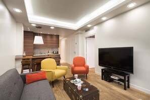 A modern living room with a grey sofa, yellow and orange chairs, a coffee table with books and a mug, a flat-screen TV, and a kitchen area with wooden cabinets and a pendant light. - 4-room apartment with 3 bedrooms in Montmartre A/C near the metro (Paris)