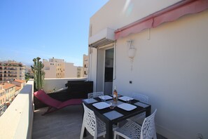 Un balcon avec une table dressée pour deux personnes, des chaises blanches, un coussin rouge et un cactus.