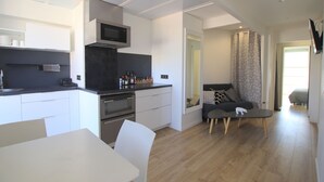 A modern kitchen with white cabinetry, a microwave, and a stove. There is a dining area with a white table and chairs. A living area with a sofa and a small table is visible. - La terrasse de Carras (Nice)