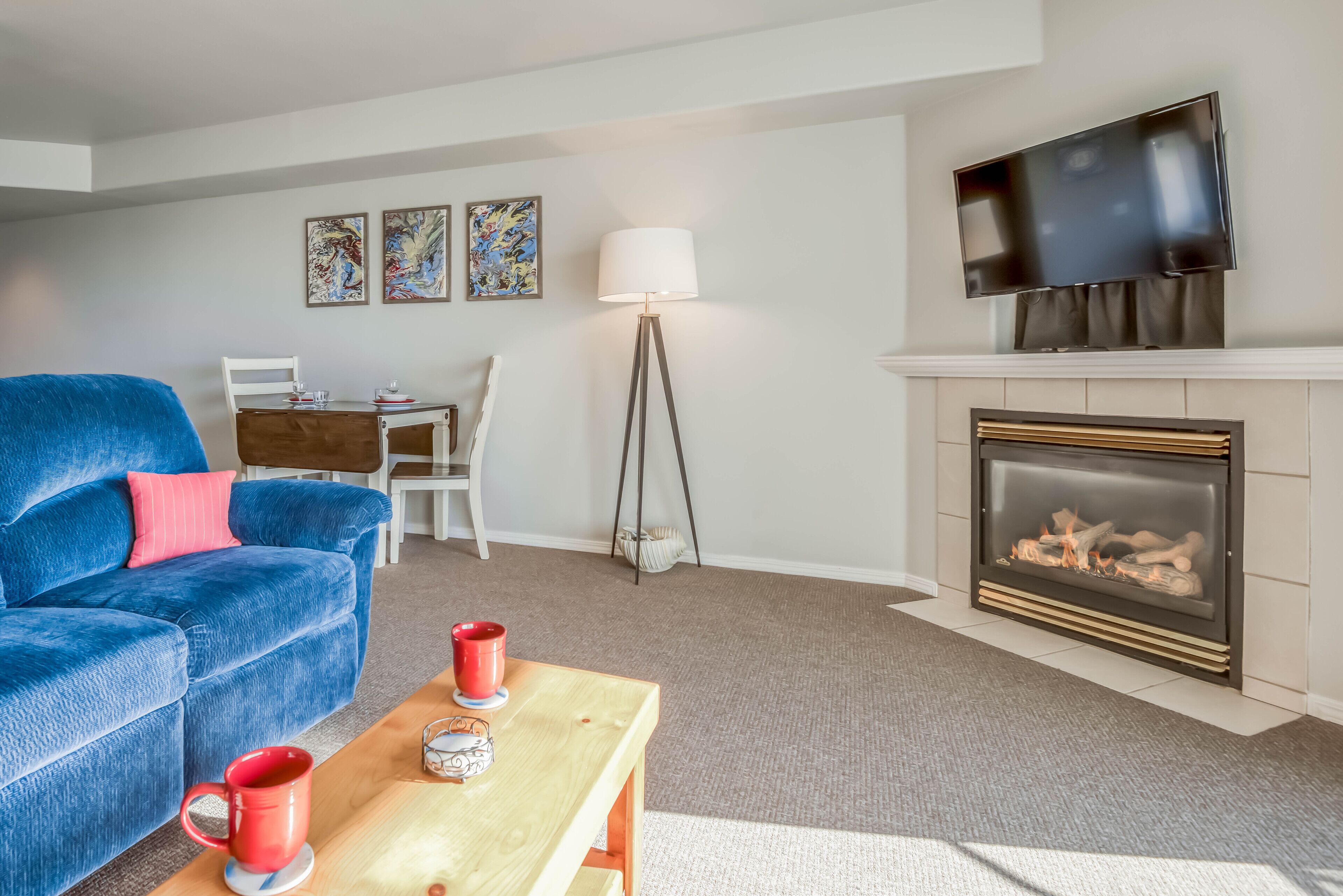 Una sala de estar con un sofá azul, una mesa de centro de madera con tazas rojas, una chimenea, un televisor montado encima y una zona de comedor con una mesa y sillas blancas.