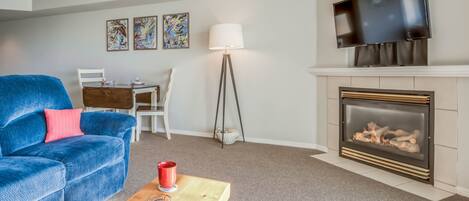 Una sala de estar con un sofá azul, una mesa de centro de madera con tazas rojas, una chimenea, un televisor montado encima y una zona de comedor con una mesa y sillas blancas.