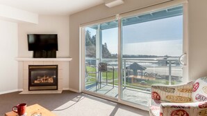 Un salón con chimenea, televisor y un gran ventanal corredero que da a un balcón con vistas a una playa y un muelle.