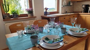 A dining table set with glassware, plates, and a bowl of greens, in a room with wooden furniture and a view of greenery outside. - Apartment "Weitblick" Greiz in the Vogtland for max. 6 people  (Greiz)
