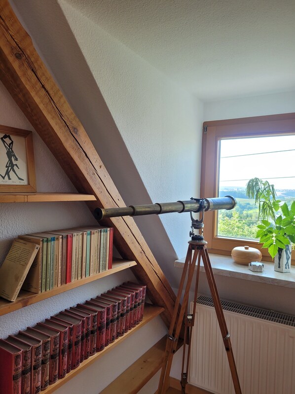 Un telescopio sobre un trípode en una habitación con vigas de madera, estanterías llenas de libros, una ventana con vistas y una planta en maceta.