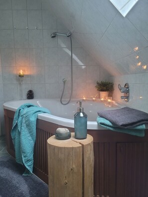 Un baño moderno con una bañera independiente, un cabezal de ducha, un taburete de madera con una botella y una piedra, y una planta en maceta.