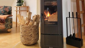 A cozy living room with a wood-burning stove, a woven basket of firewood, a wooden ladder, and a framed picture on the wall. - Apartment "Weitblick" Greiz in the Vogtland for max. 6 people  (Greiz)