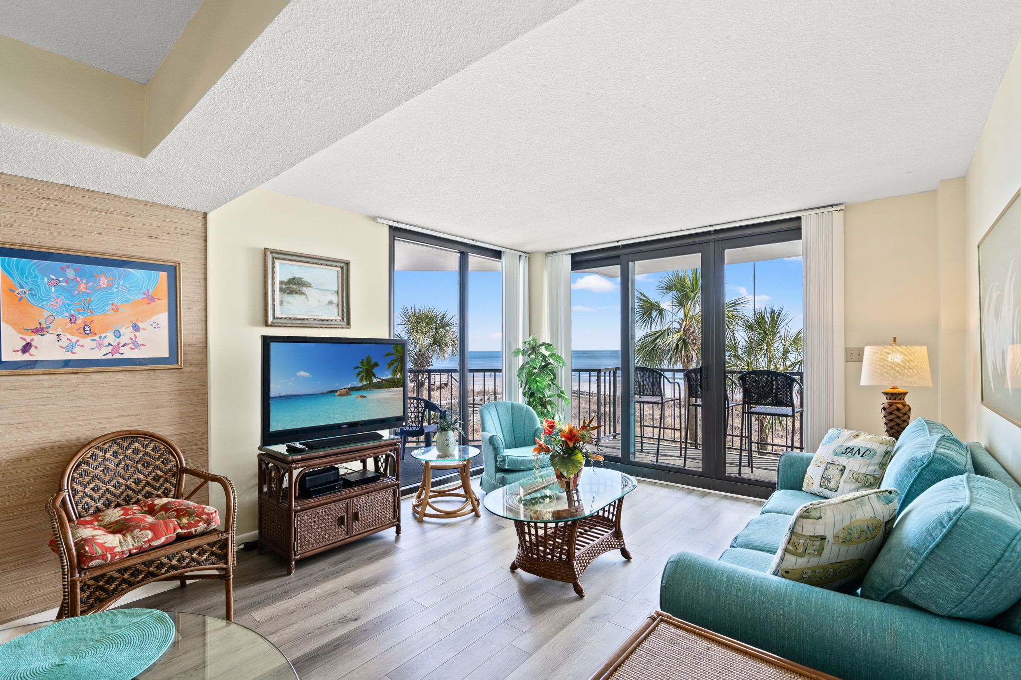 A spacious living area with a large TV, comfortable seating, a glass coffee table, and a balcony with a view of the ocean.