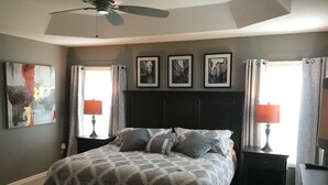 Un dormitorio con una cama grande, dos mesitas de noche con lámparas, un ventilador de techo y tres cuadros en la pared.