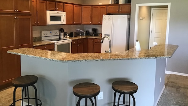 Una cocina con armarios de madera, encimera de granito, microondas, refrigerador, horno, lavamanos y tres taburetes.