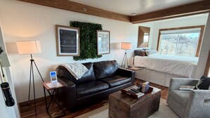 A modern living room with a black leather sofa, a wooden coffee table, and a grey armchair. There are two framed artworks on the wall, a window with a view of trees, and a lamp on a tripod stand. - The Hideaway at Oma's Meadow (Sugar Mountain)