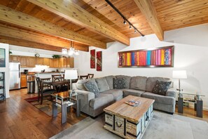 Un espacio habitable moderno con techo de madera, un sofá seccional gris, una mesa de centro de vidrio y una zona de cocina con armarios de madera.