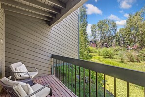 A balcony with a wooden deck, two chairs, and a view of a green landscape with trees and a building in the distance. - Riverside Dream | Short Walk to Main Street & Hike (Telluride)