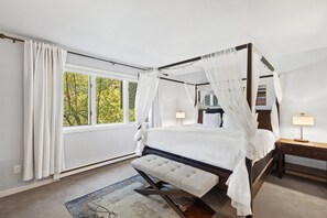 Uma cama com dossel e cortina, uma mesa de cabeceira com abajur e um banco ao pé da cama.