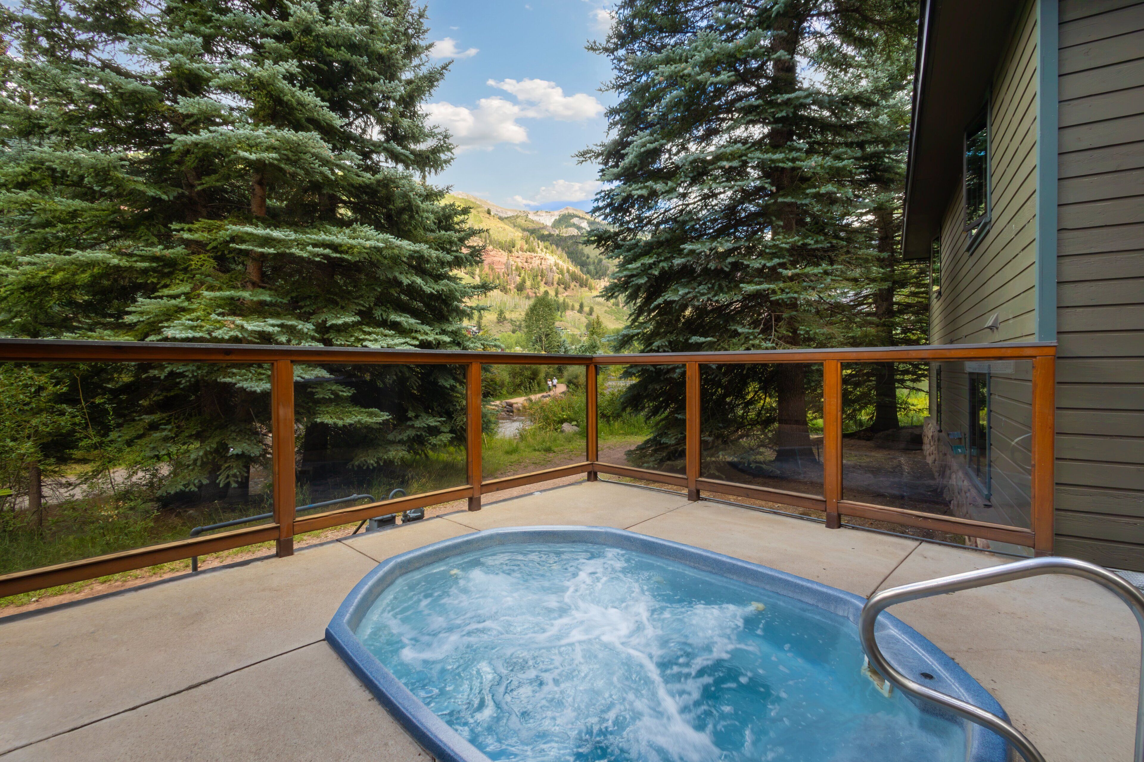 A hot tub with a wooden deck and glass railing, surrounded by trees and a view of a valley.
