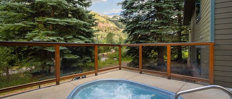 A hot tub with a wooden deck and glass railing, surrounded by trees and a view of a valley.