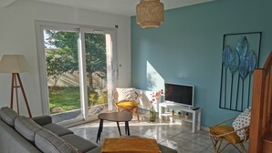 A living room with a sofa, a TV on a white stand, a wicker chair, and a large window with a view of greenery. - Maison de Vacances Moguériec (Sibiril)