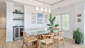 A dining area with a glass table, wooden chairs, and a chandelier. There is a refrigerator and a plant in the background.