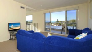 A living room with a TV on a stand, a blue sofa, and a balcony with a view of the sea.