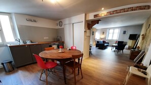 A spacious living area with a wooden dining table, red chairs, a kitchen with a sink and cabinets, and a sofa in the background. - Apartment (Guérande)