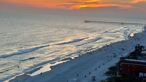 Una spiaggia con un molo che si protende nell'acqua, persone che passeggiano sulla riva e un tramonto sullo sfondo.