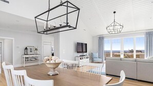 A modern living room with a large window, a dining area with a wooden table and white chairs, and a gray sofa.