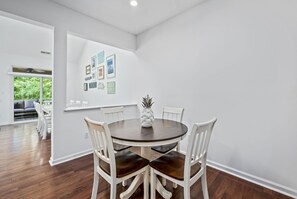 Un comedor con una mesa redonda, sillas blancas y un centro de mesa con piña. Hay cuadros en la pared y se ve un balcón con un sofá.