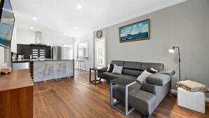Ein modernes Wohnzimmer mit einem grauen Sofa, einem Couchtisch und einem Fernseher. Es gibt eine Küchenzeile mit Kühlschrank, Mikrowelle und Barhocker. An der Wand hängt ein Gemälde eines Schiffes, und eine Uhr ist ebenfalls zu sehen.