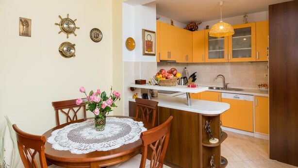 A compact kitchen with orange cabinets, a white countertop, and a small island with a sink. There is a dining table with chairs and a vase of flowers. - Apartment Jele (Zupa dubrovacka)