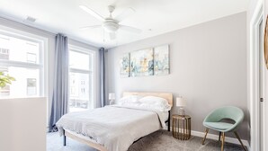 Ein Schlafzimmer mit einem Bett, einem Deckenventilator, einem Fenster mit Vorhängen, einem Nachttisch mit Lampe und einem Sessel.