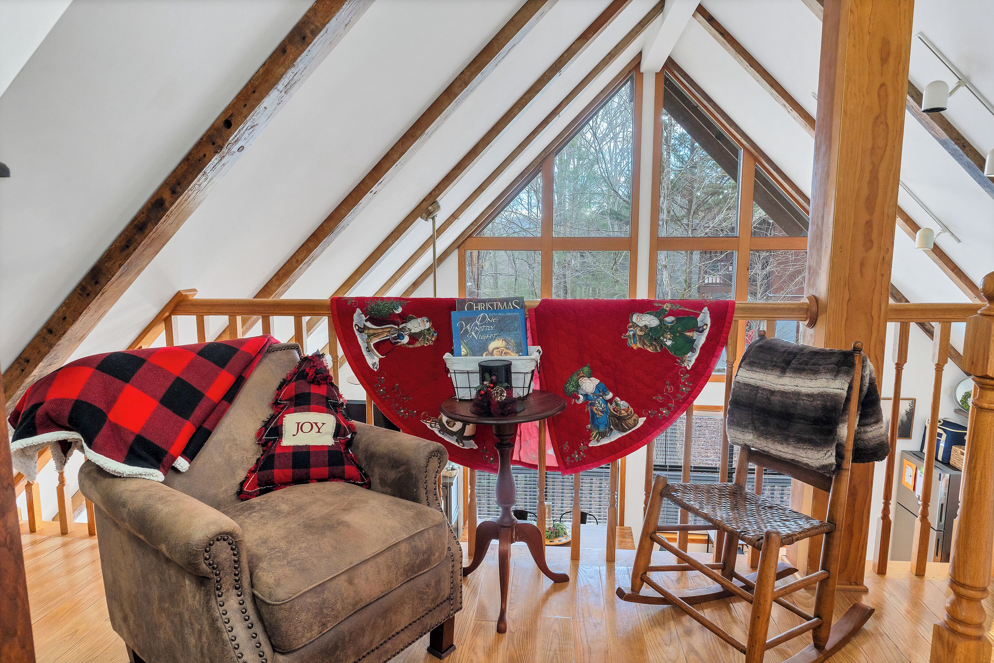 Een knus loft slaapkamer met een schuine plafond, houten balken, een groot raam, een zwart-rood geruit plaid op een stoel en een kersttafelkleed.