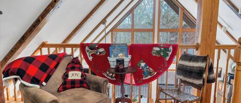 Een knus loft slaapkamer met een schuine plafond, houten balken, een groot raam, een zwart-rood geruit plaid op een stoel en een kersttafelkleed.