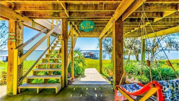 A wooden staircase leading to a covered area with a 'hello' sign, a swing, and a view of a house across a water body.
