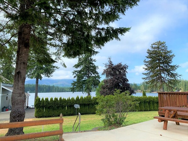 A lakeside patio with a wooden bench and picnic table, surrounded by trees and a view of the lake.