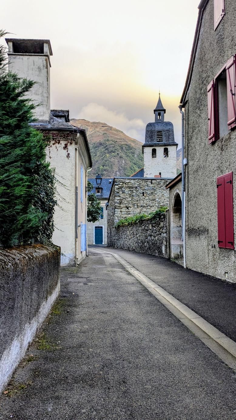 Une rue étroite bordée d’immeubles en pierre, un clocher d’église et une montagne en arrière-plan.