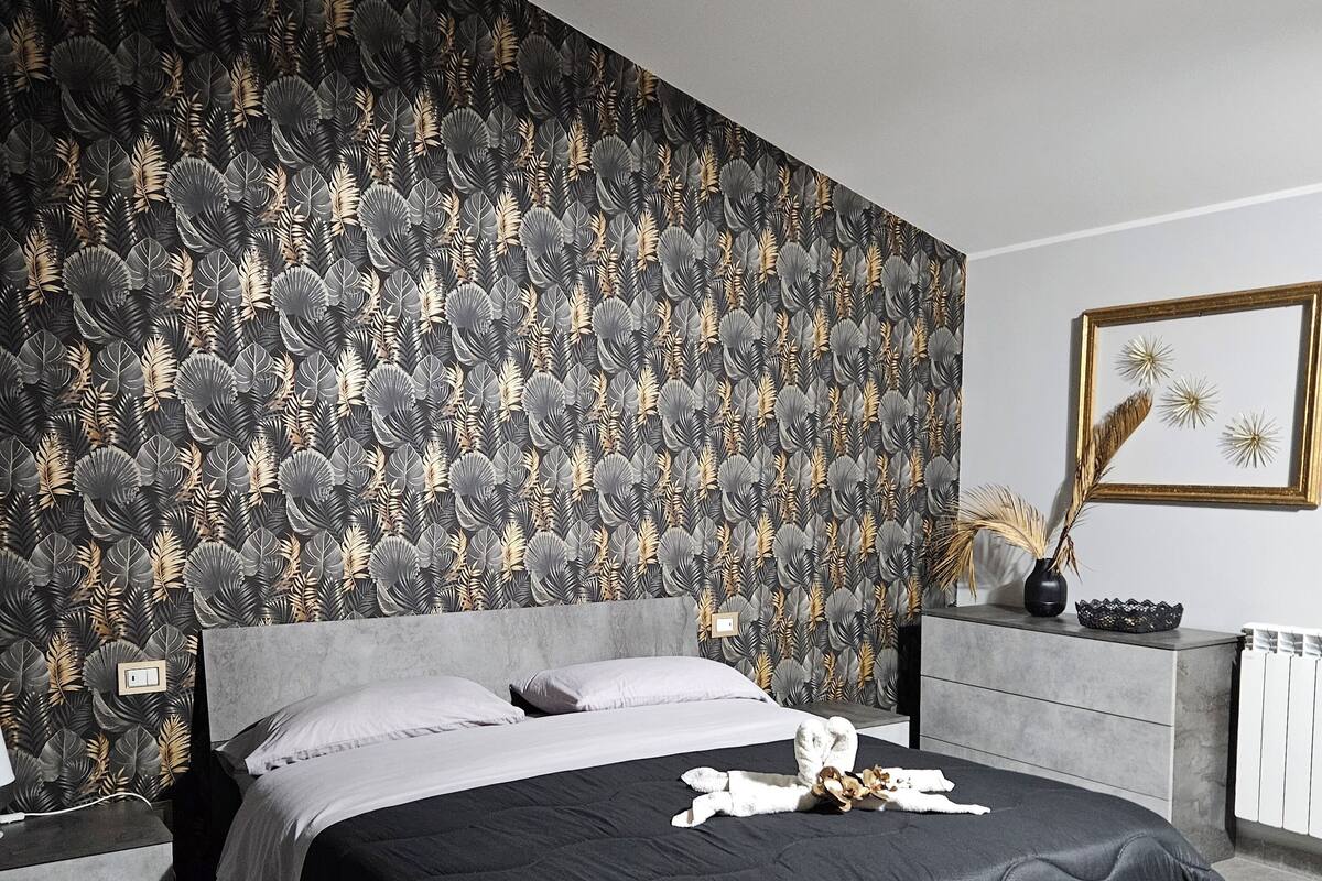 Une chambre moderne avec un grand lit, un papier peint à motifs gris et noir, une table de chevet et un tableau encadré au mur.